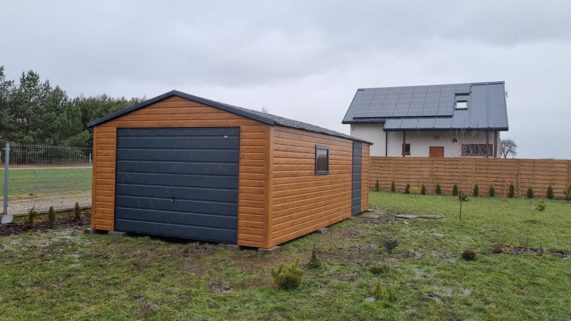 Drewnopodobny garaż z szarą bramą segmentową i małym oknem, ustawiony na betonowych bloczkach na trawniku, w tle dom z panelami słonecznymi i drewniany płot.