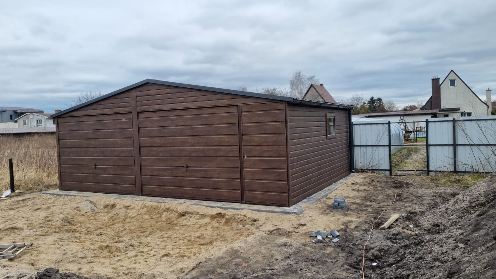 Brązowy, dwustanowiskowy garaż blaszany z oknem bocznym, posadowiony na fundamencie z kostki, widok z boku na tle działki w trakcie prac ziemnych.
