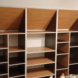 Półki regału z białymi ramami i drewnianym wypełnieniem, część z nich jest pusta, a część ma drewniane panele na tylnej ścianie, widoczne materiały budowlane na blacie.