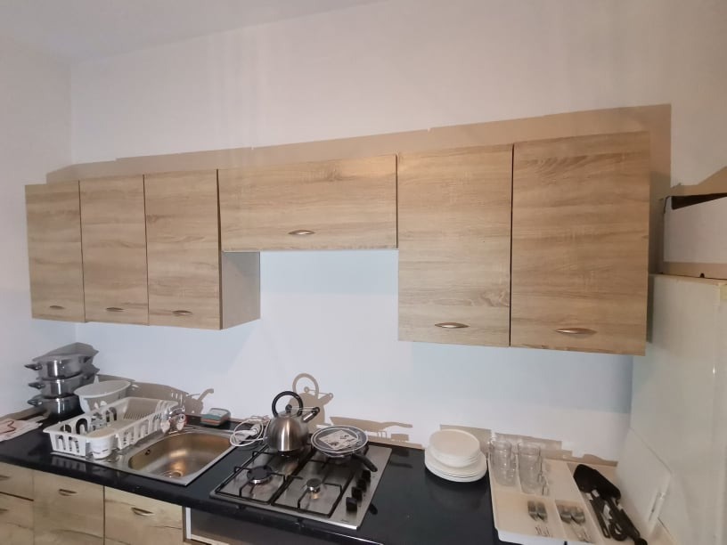 Jasne, drewnopodobne szafki kuchenne wiszące nad czarnym blatem z kuchenką gazową i zlewozmywakiem. Na blacie naczynia i akcesoria kuchenne.