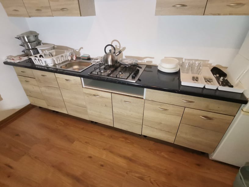 Kuchenna zabudowa z frontami imitującymi jasne drewno, czarny blat, widoczny zlew, płyta gazowa, naczynia i sztućce.