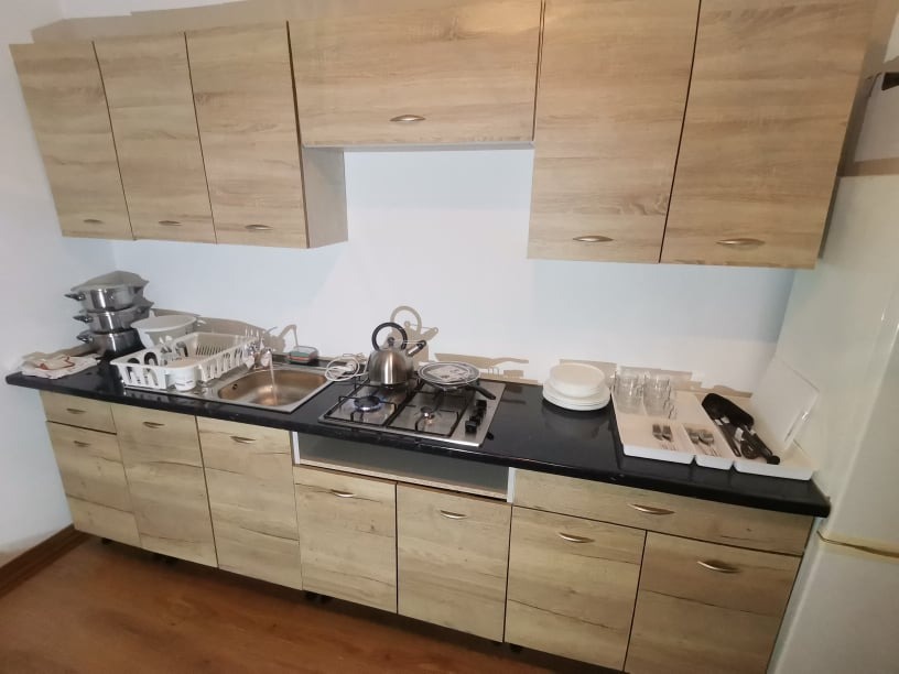 Jasna kuchnia z drewnopodobnymi frontami szafek, czarnym blatem, zlewozmywakiem i płytą gazową, naczynia na suszarce i na blacie.