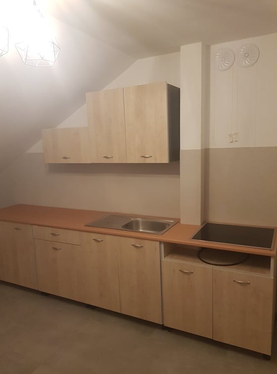 Jasna, drewnopodobna zabudowa kuchenna pod skosem dachu z pojedynczym zlewem i płytą indukcyjną, oświetlona lampą wiszącą.