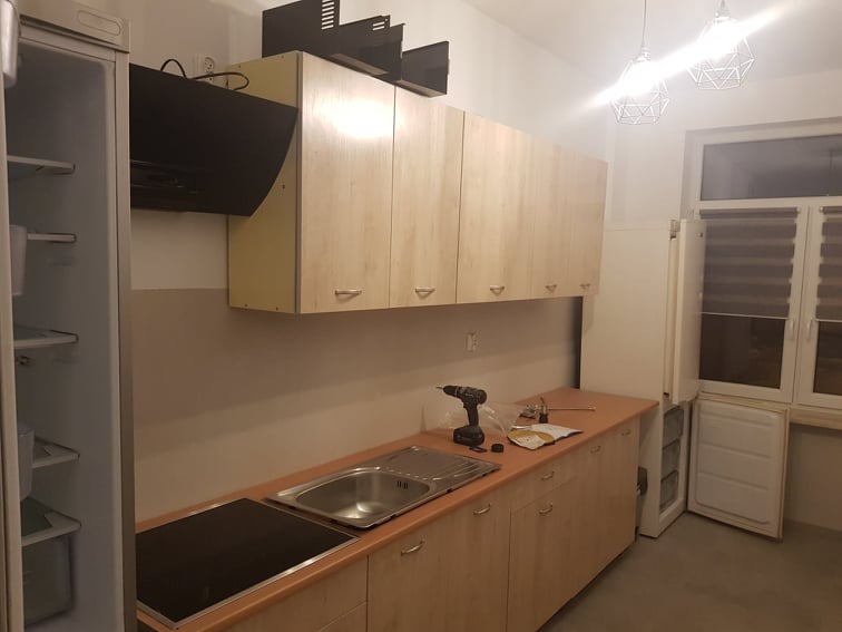 Montaż kuchni: widok na szafki wiszące z jasnego drewna, okap, zlew i płytę grzewczą. Na blacie leżą narzędzia, w tym wiertarka.