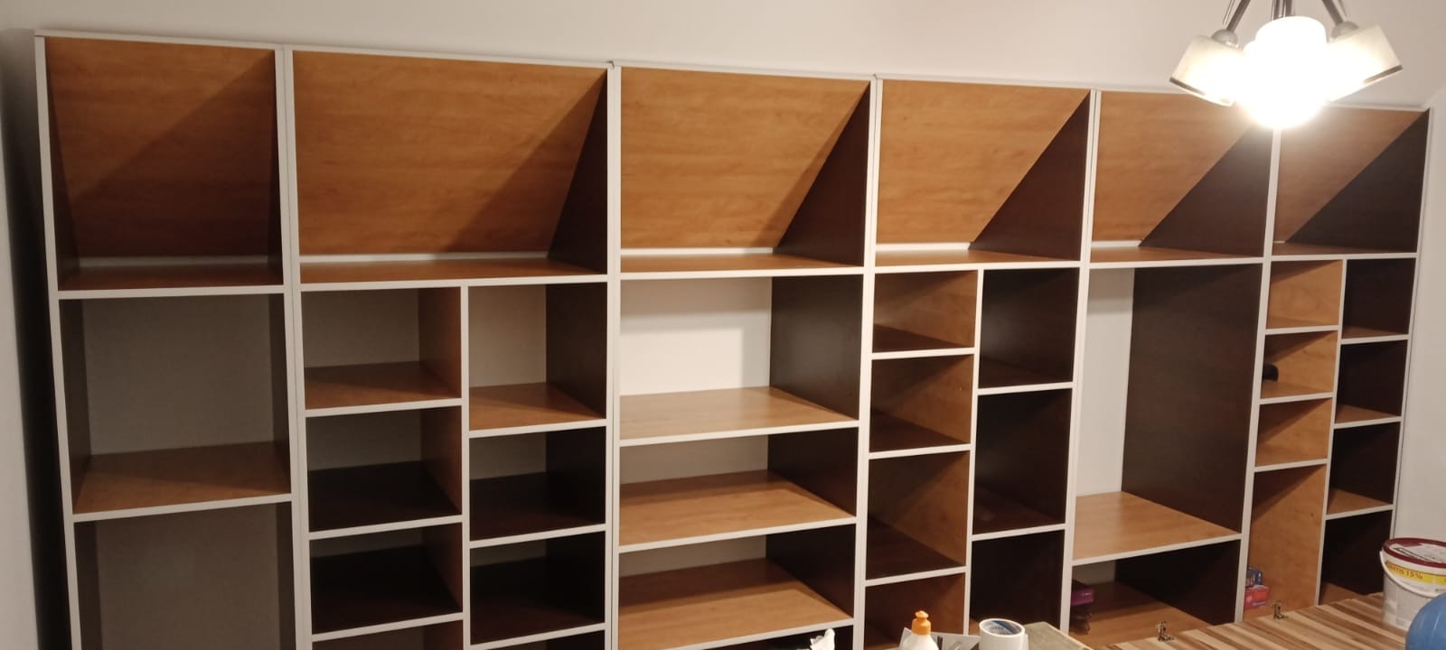 Półki regału z białymi ramami i drewnianym wypełnieniem, część z nich jest pusta, a część ma drewniane panele na tylnej ścianie, widoczne materiały budowlane na blacie.