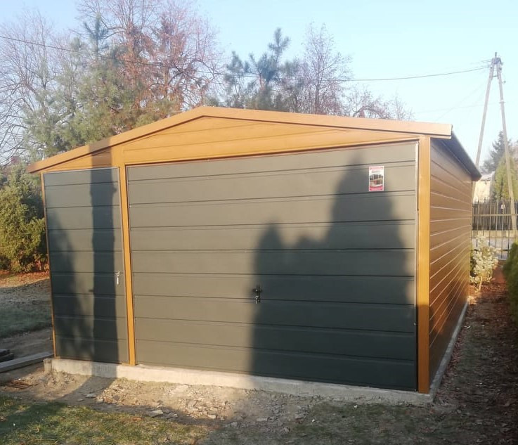 Szary blaszak z dwuspadowym dachem w kolorze drewna, z dodatkowymi drzwiami z boku, na betonowej wylewce, w tle drzewa bez liści.