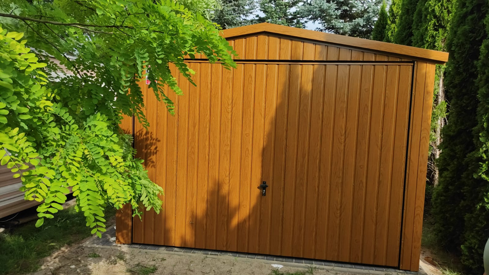Brązowy garaż blaszany z pionowym wzorem desek, częściowo zasłonięty przez gałąź drzewa, z ciemnym dachem i czarnym zamkiem.