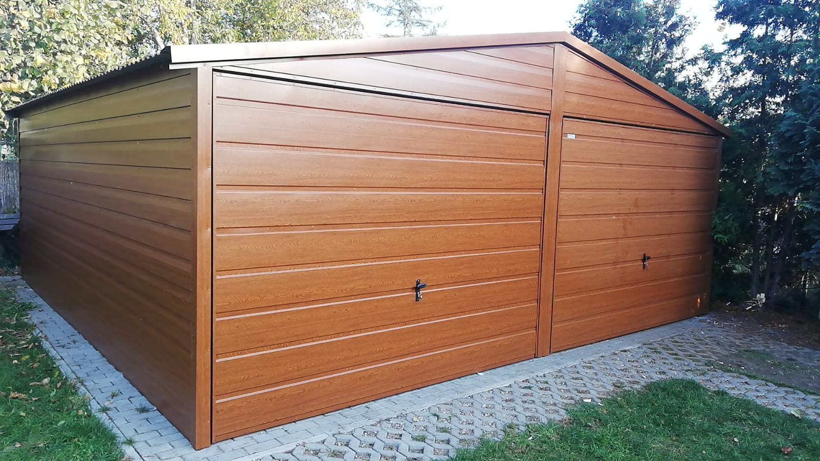 Brązowy, dwustanowiskowy garaż blaszany z poziomymi panelami, usytuowany na kostce brukowej, otoczony zielenią.