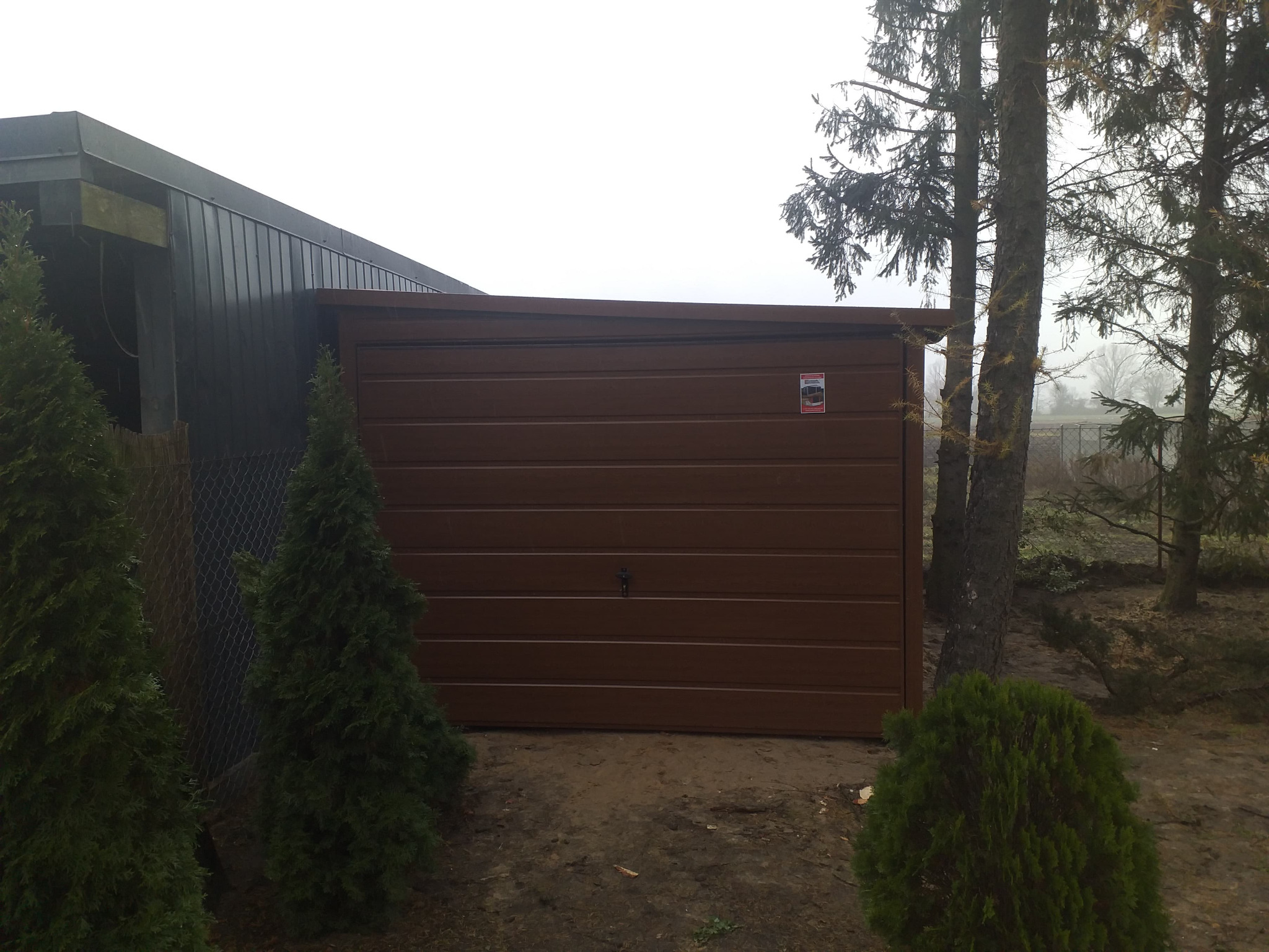 Brązowy garaż blaszany z poziomymi przetłoczeniami, przylegający do ciemnego budynku, otoczony zielonymi tujami i drzewami w pochmurny dzień.
