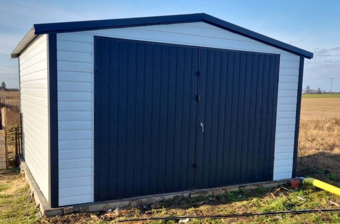 Gotowy, biało-grafitowy garaż z blachy falistej, z dwuskrzydłowymi drzwiami, na podmurówce, na tle zaoranego pola i błękitnego nieba.