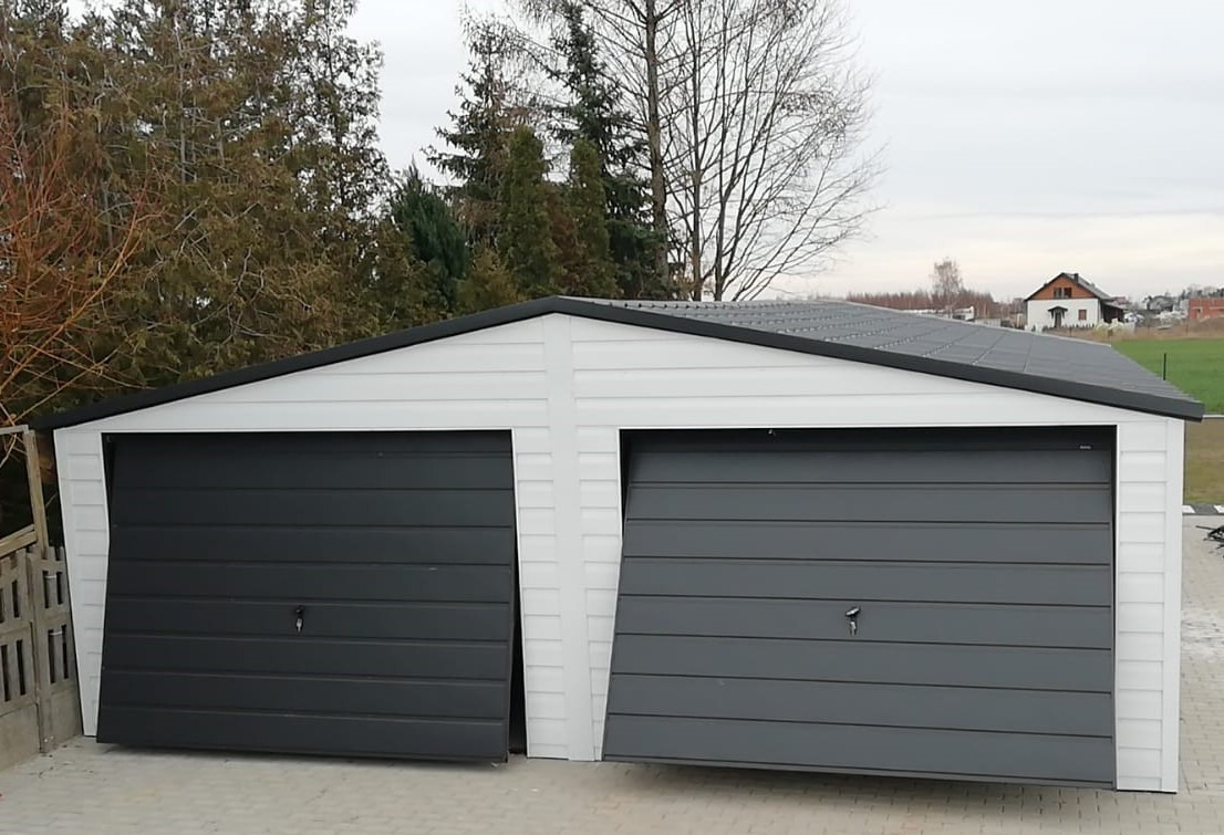 Dwustanowiskowy garaż blaszany z szarymi bramami segmentowymi, jedna brama lekko uchylona, białe ściany z panelami, czarny dach, widok z zewnątrz na tle drzew i domu.