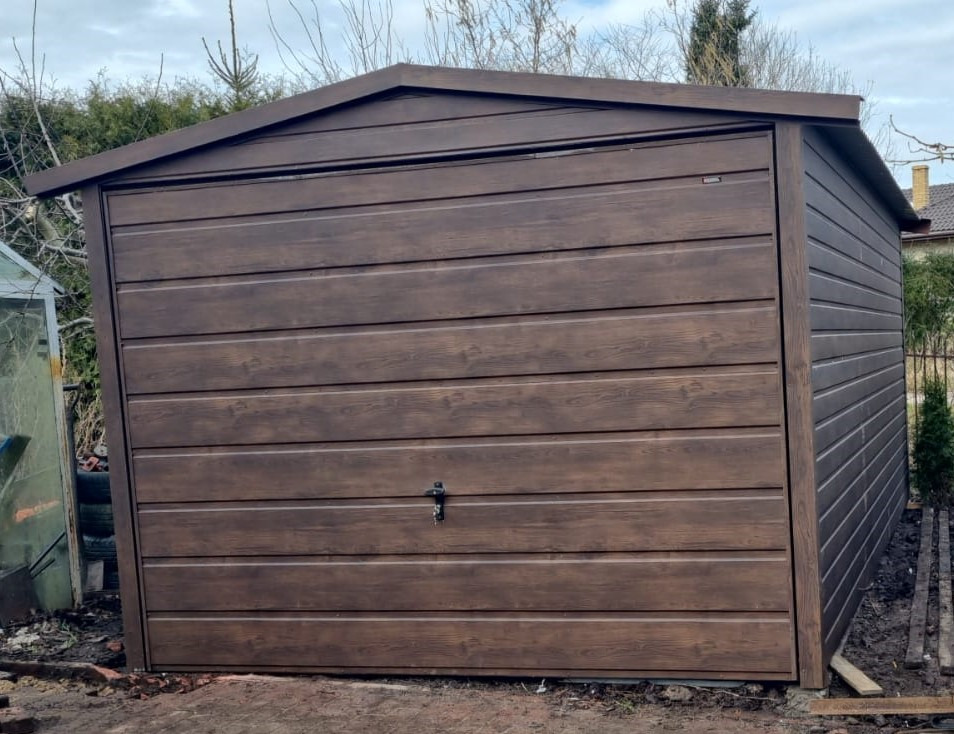 Nowoczesny, brązowy garaż blaszany z imitacją drewna, zamykany na klucz, ustawiony na nierównym, ziemistym podłożu z widocznymi elementami starej kostki brukowej.