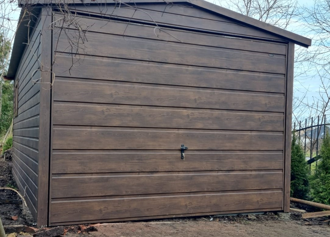 Brązowy garaż blaszany z poziomymi panelami imitującymi drewno, widoczny z zewnątrz na posesji z ogrodzeniem i zielenią w tle.