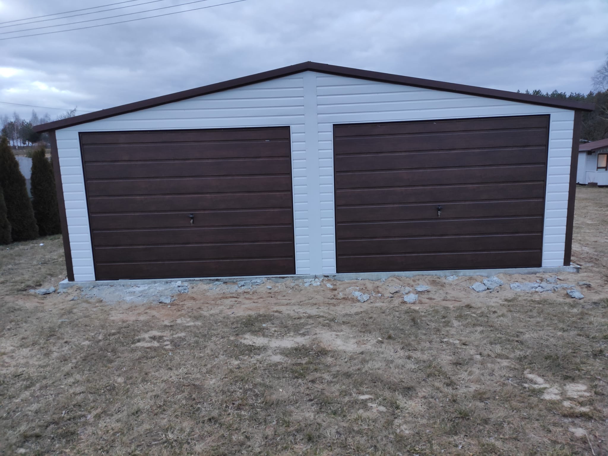 Dwustanowiskowy garaż blaszany z brązowymi bramami segmentowymi i białymi ścianami, świeżo posadowiony na fundamencie, widoczne resztki gruzu.