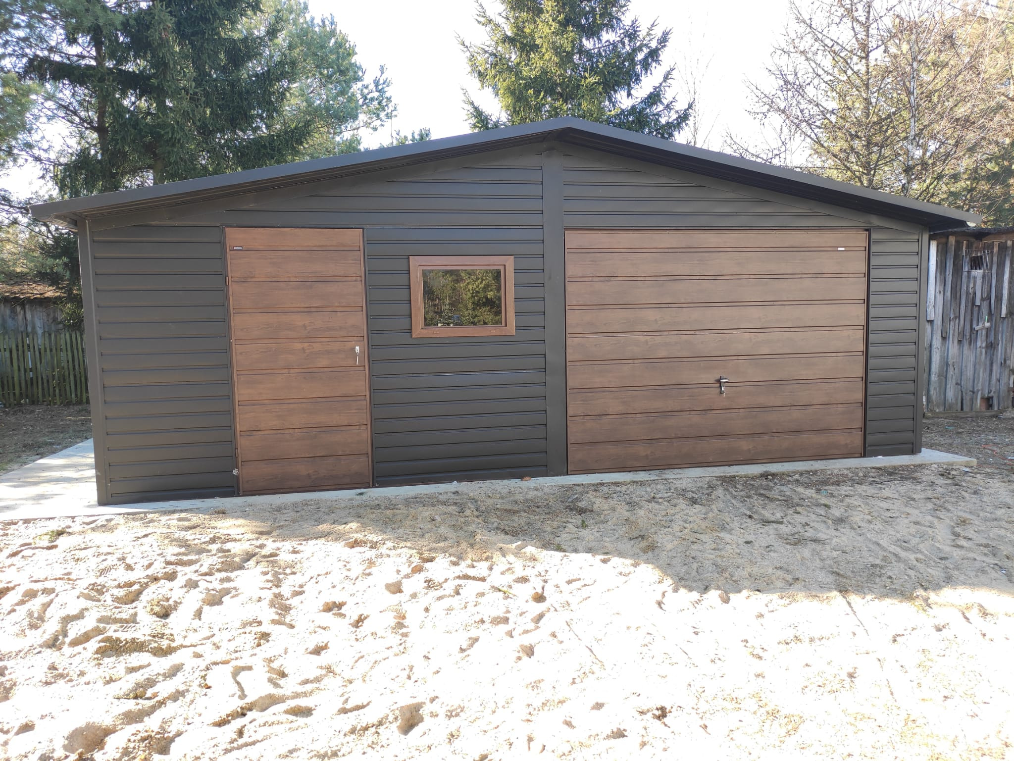 Gotowy, dwustanowiskowy garaż blaszany z brązowymi drzwiami segmentowymi, oknem i grafitową blachą trapezową, otoczony piaszczystym terenem.