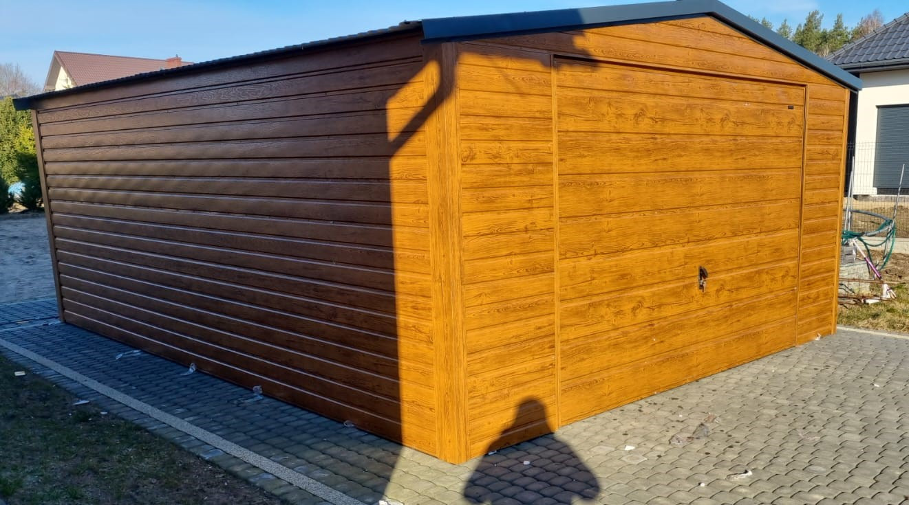 Zmontowany, brązowy garaż z blachy imitującej drewno, z czarnym dachem, ustawiony na kostce brukowej w słoneczny dzień, cień osoby w rogu.