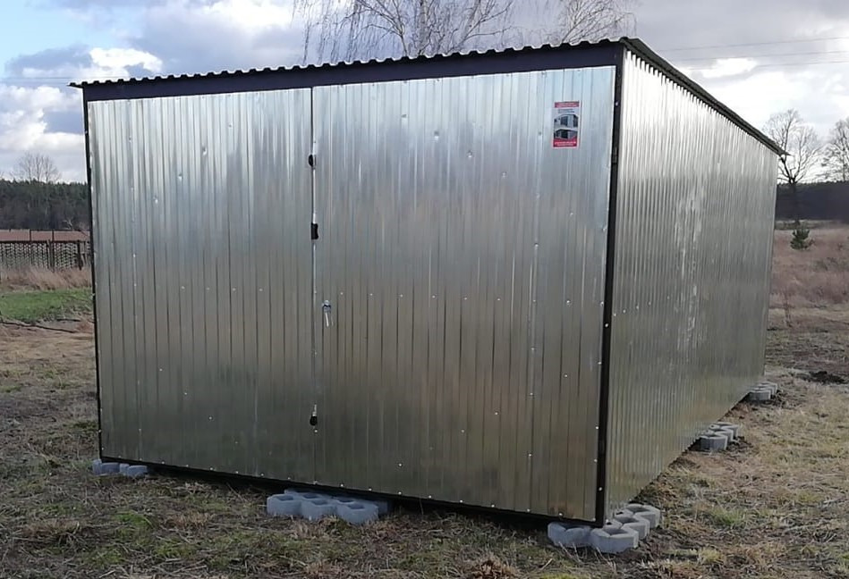 Metalowy garaż z dwuskrzydłowymi drzwiami, ustawiony na betonowych bloczkach, z naklejoną etykietą producenta na ścianie