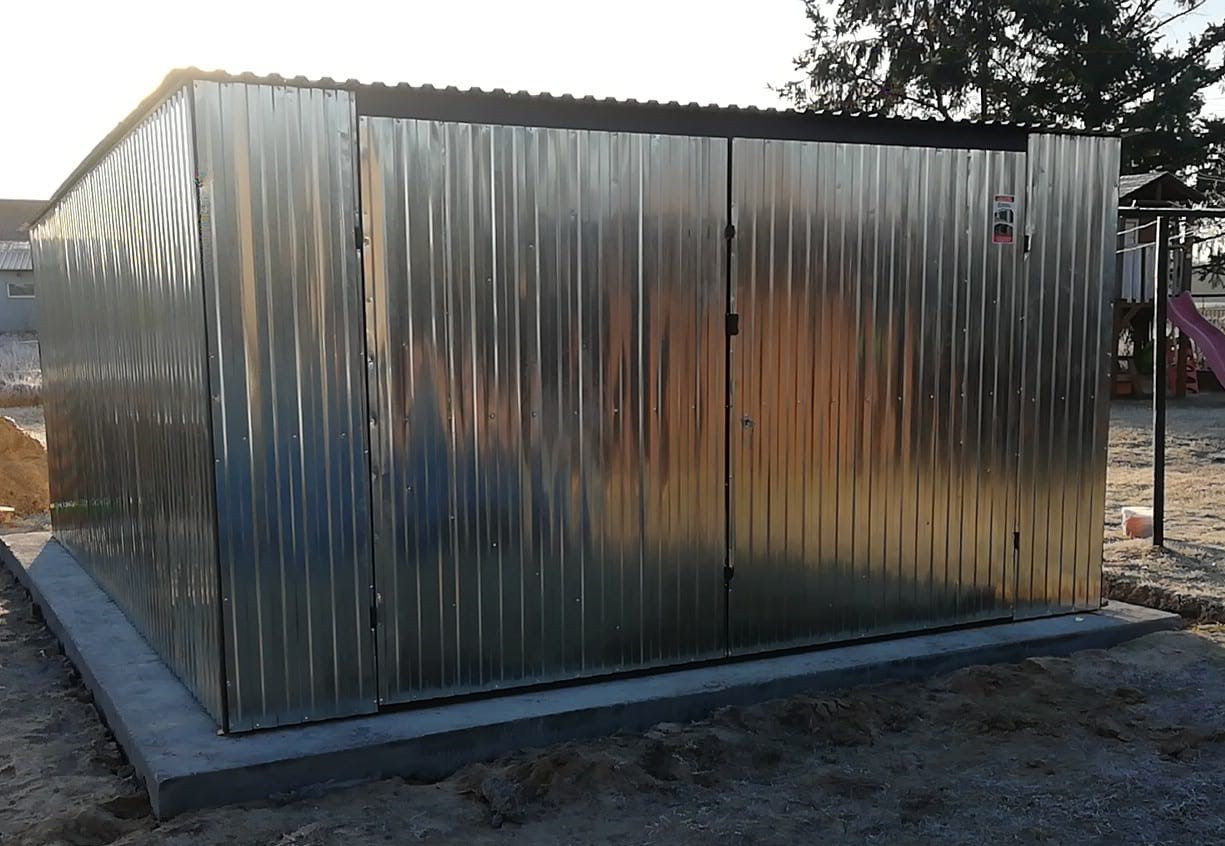 Metalowy garaż z blachy falistej, posadowiony na betonowej wylewce, widoczny w tle plac zabaw.