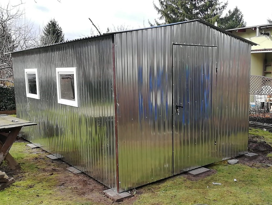 Metalowy budynek gospodarczy z dwoma oknami i drzwiami, ustawiony na betonowych płytach w ogrodzie.