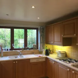 Jasna kuchnia z drewnianymi szafkami, okno z widokiem na ogród, żółte płytki nad blatem i sprzęt AGD.