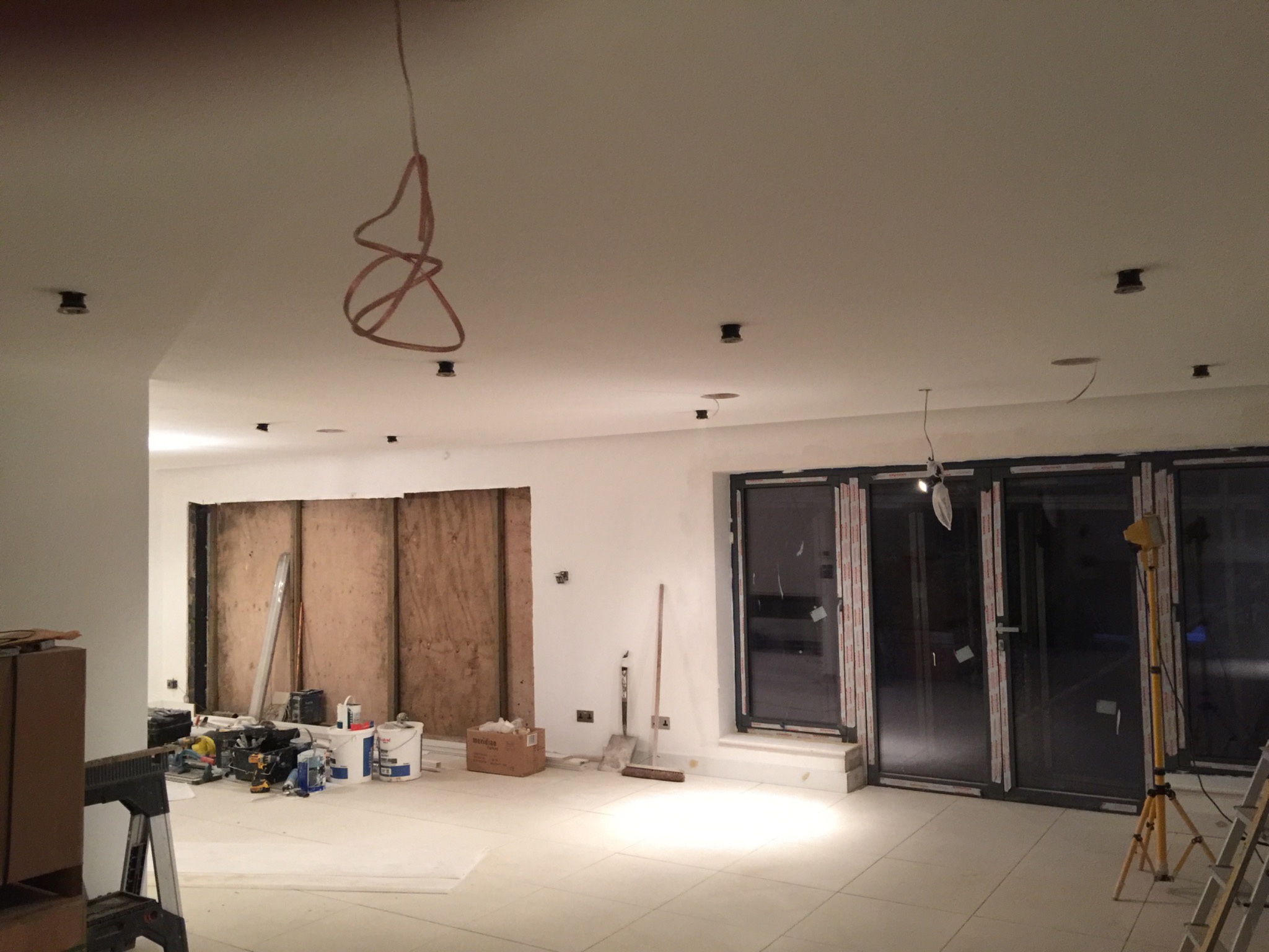 Wnętrze w trakcie prac wykończeniowych, z widocznymi narzędziami, materiałami budowlanymi, odsłoniętymi przewodami pod sufitem i dużymi, przesuwnymi drzwiami balkonowymi