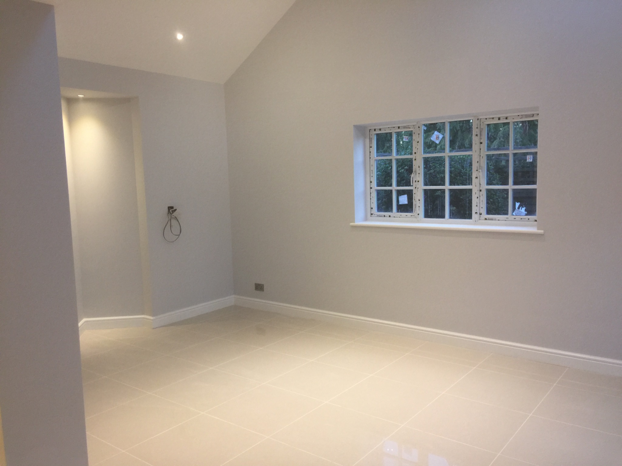 Jasne, puste pomieszczenie z kremowymi płytkami podłogowymi, świeżo pomalowanymi szarymi ścianami, białymi listwami przypodłogowymi i oknem z widokiem na zieleń. Wnęka z oświetleniem punktowym po...