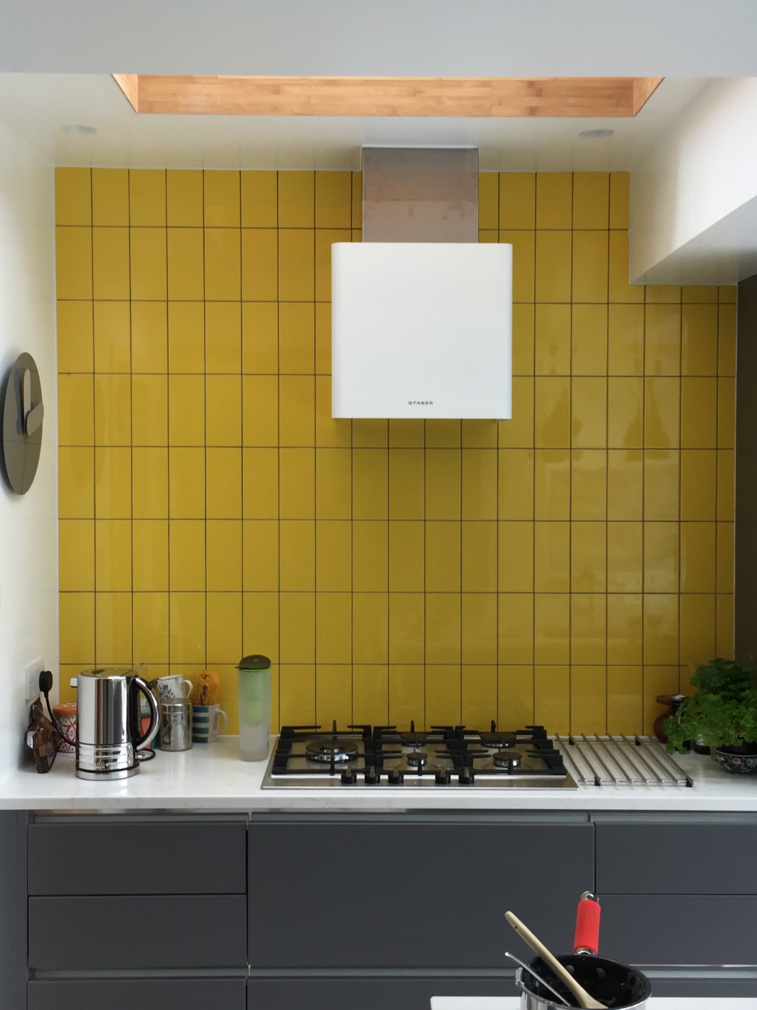 Żółte płytki na ścianie w kuchni, nad blatem z szarymi szafkami, biały okap Faber, czarny zegar na ścianie po lewej stronie.