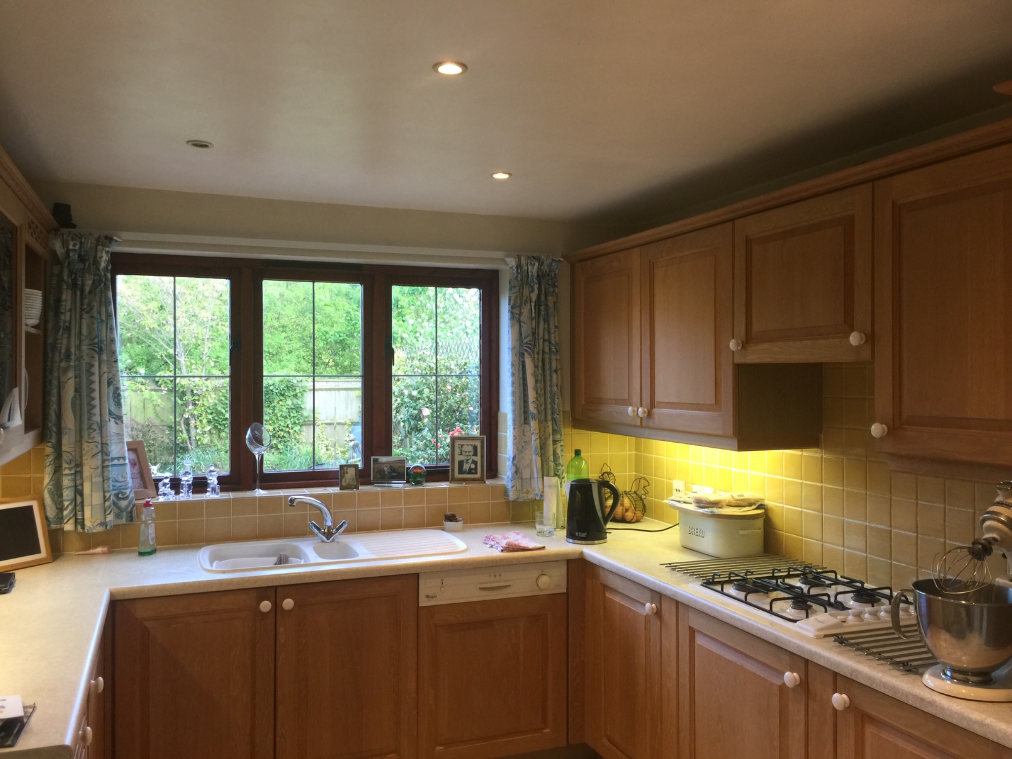 Jasna kuchnia z drewnianymi szafkami, okno z widokiem na ogród, żółte płytki nad blatem i sprzęt AGD.