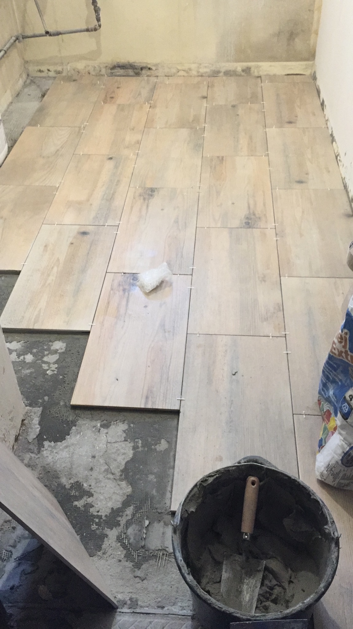 Układanie podłogi z płytek imitujących drewno, widoczny fragment podłogi z ułożonymi płytkami, krzyżyki dystansowe, wiadro z zaprawą i kielnią, oraz nieułożone płytki.