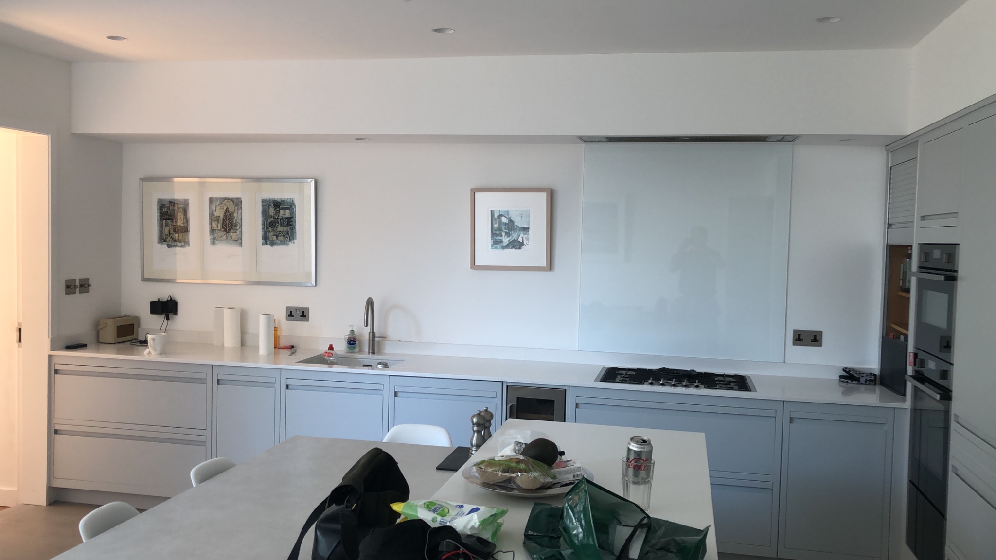 Jasna, nowoczesna kuchnia z szarymi szafkami, białym blatem i wyspą, ozdobiona obrazami na ścianie i wyposażona w sprzęt AGD, w tle odbicie osoby w szklanym panelu.