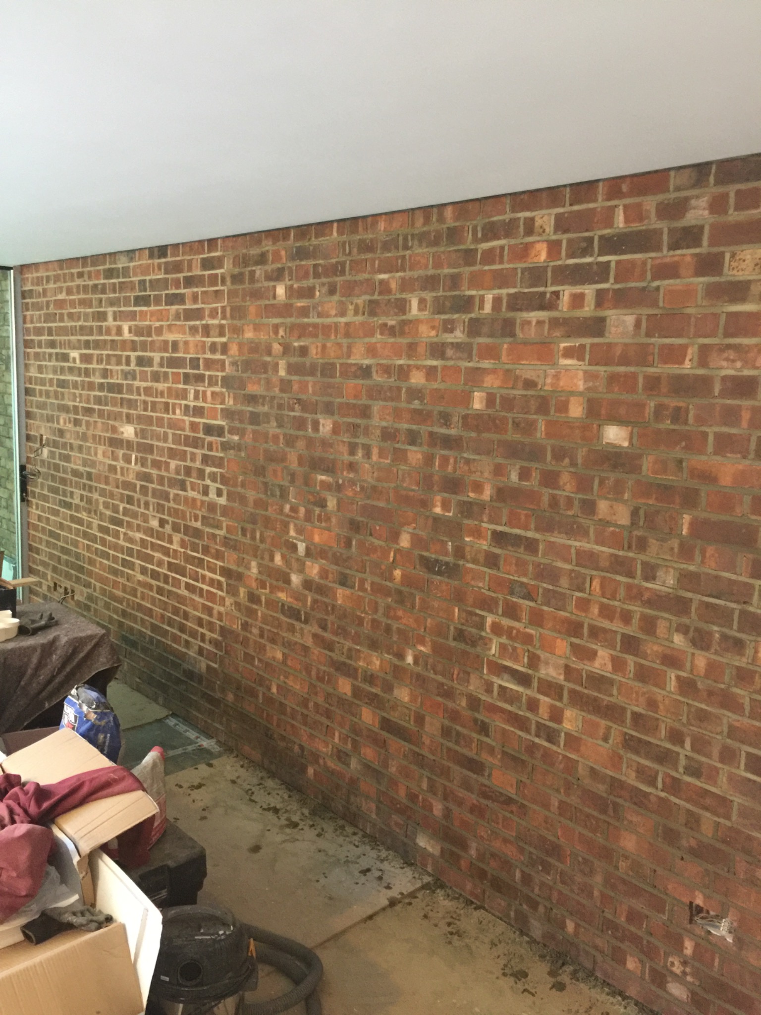 Ceglana ściana wewnętrzna w trakcie remontu, widoczne ślady po pracach na podłodze i częściowo zakryta podłoga, biały sufit.