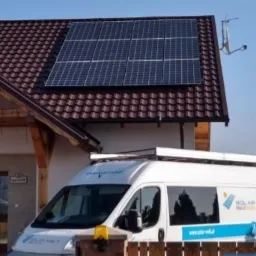 Dom jednorodzinny z brązowym dachem, na którym zamontowano panele fotowoltaiczne; na podjeździe stoi biały samochód dostawczy z logo firmy instalującej fotowoltaikę.