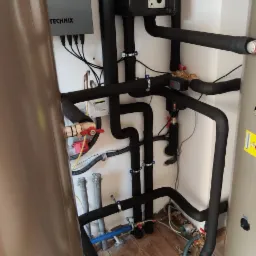 Instalacja grzewcza w kotłowni z widocznym zbiornikiem buforowym, rurami w izolacji, zaworami i panelem sterowania TECHNiX na ścianie.