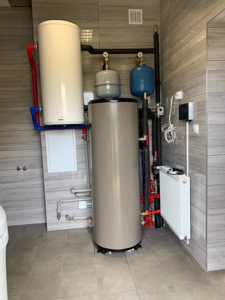 Kaskadowy układ dwóch zbiorników akumulacyjnych, z szarym zbiornikiem na dole i białym na górze, połączonych rurami w kolorach czerwonym i niebieskim, z dodatkowym małym grzejnikiem panelowym...