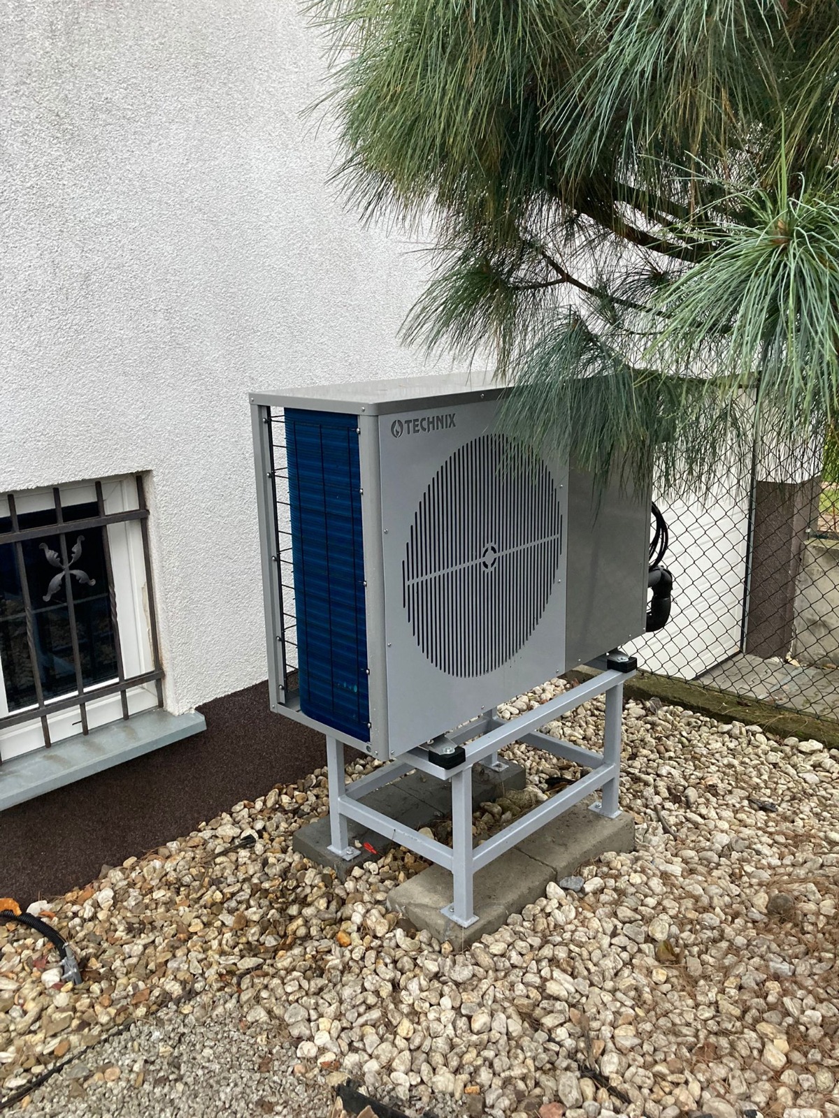Szara jednostka zewnętrzna z niebieskimi lamelami marki Technix, zamontowana na metalowym stelażu na żwirowym podłożu, z widocznym oknem piwnicznym i gałęziami iglastego drzewa.