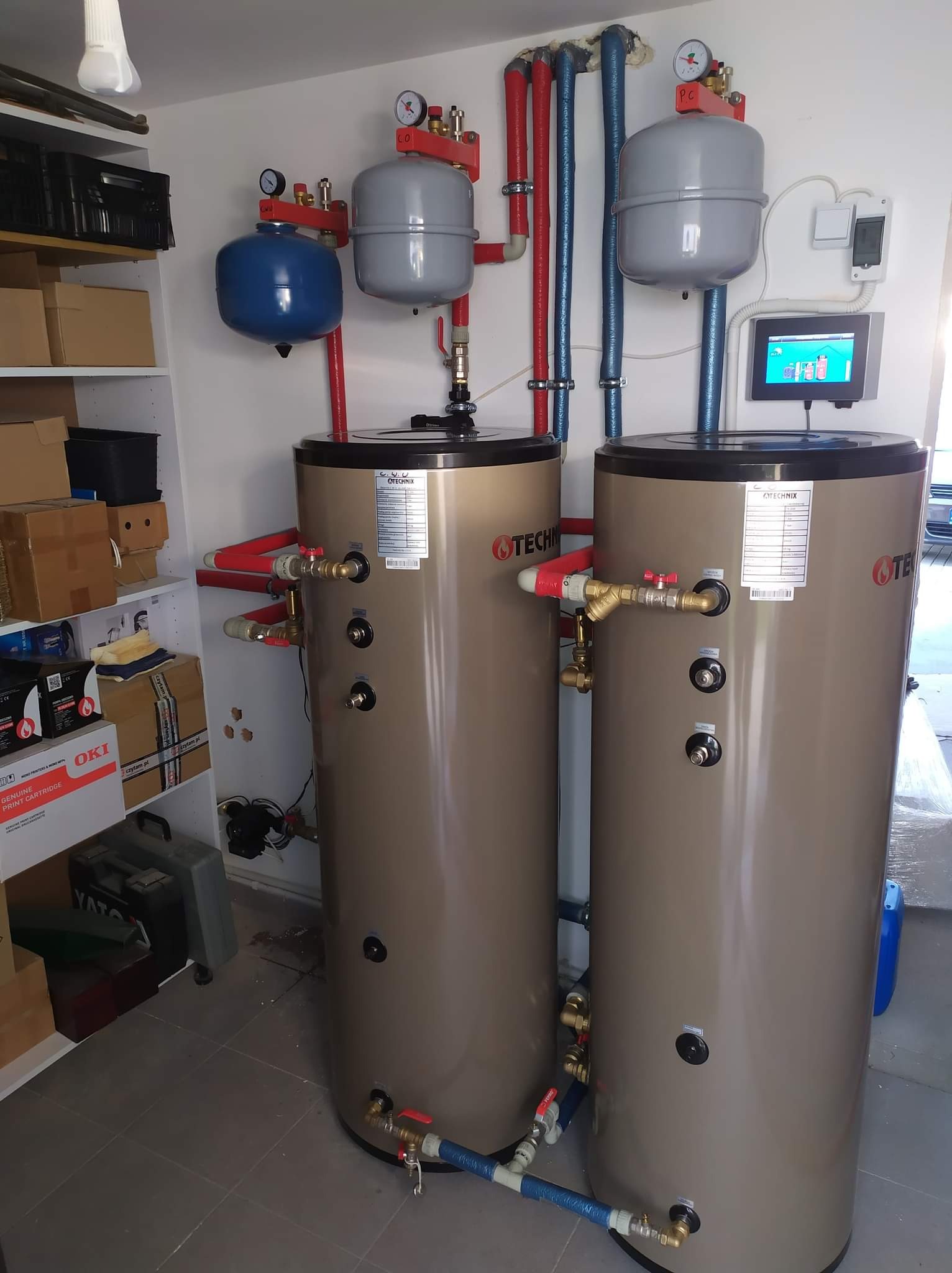 Dwa brązowe zasobniki ciepłej wody użytkowej marki TECHNIX w kotłowni, z podłączonymi rurami w kolorze czerwonym i niebieskim, manometry na górze i ekran sterujący na ścianie.