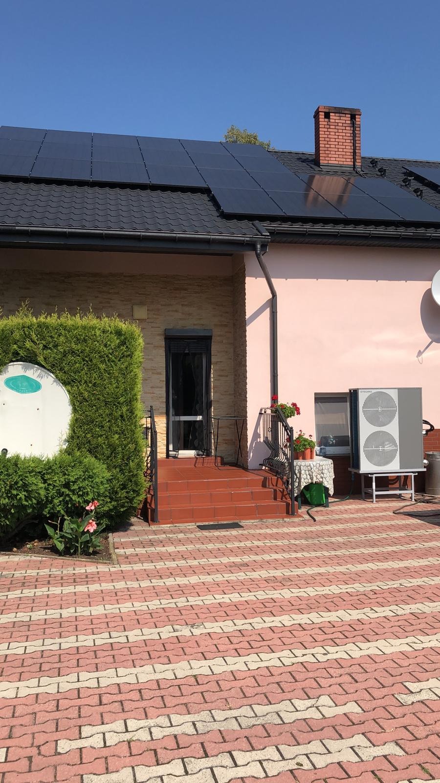 Dom jednorodzinny z panelami słonecznymi na dachu i jednostką zewnętrzną systemu grzewczego na posesji, widok z poziomu brukowanego podjazdu.