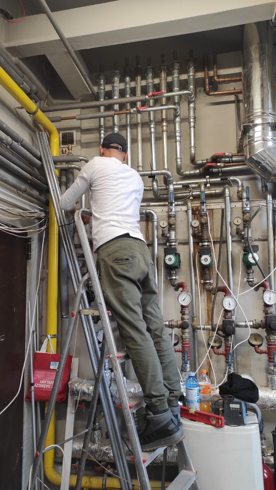 Instalator na drabinie, wykonujący prace przy rozbudowanej instalacji grzewczej z licznymi rurami, zaworami i manometrami, butelki wody i poziomica na obudowie urządzenia.