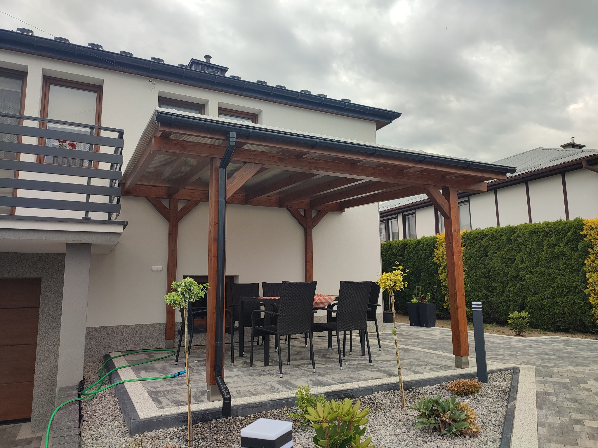 Zadaszony taras z drewnianą konstrukcją i poliwęglanowym dachem, wyposażony w czarne krzesła i stół nakryty kratkowanym obrusem, otoczony ozdobnymi krzewami i kamieniami.