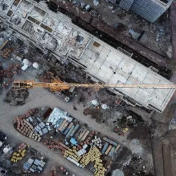 Widok z drona na plac budowy z kilkoma budynkami w stanie surowym, dźwigiem i stertami materiałów budowlanych w różnych kolorach.