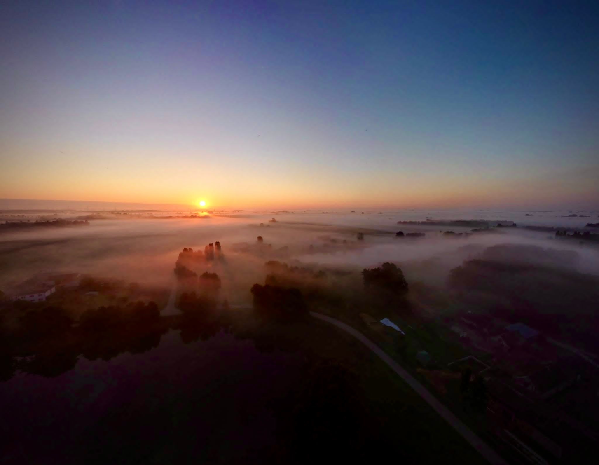 Panoramiczny widok z drona na wschód słońca nad mglistą doliną, promienie słoneczne przebijają się przez drzewa, widoczne domy i droga.