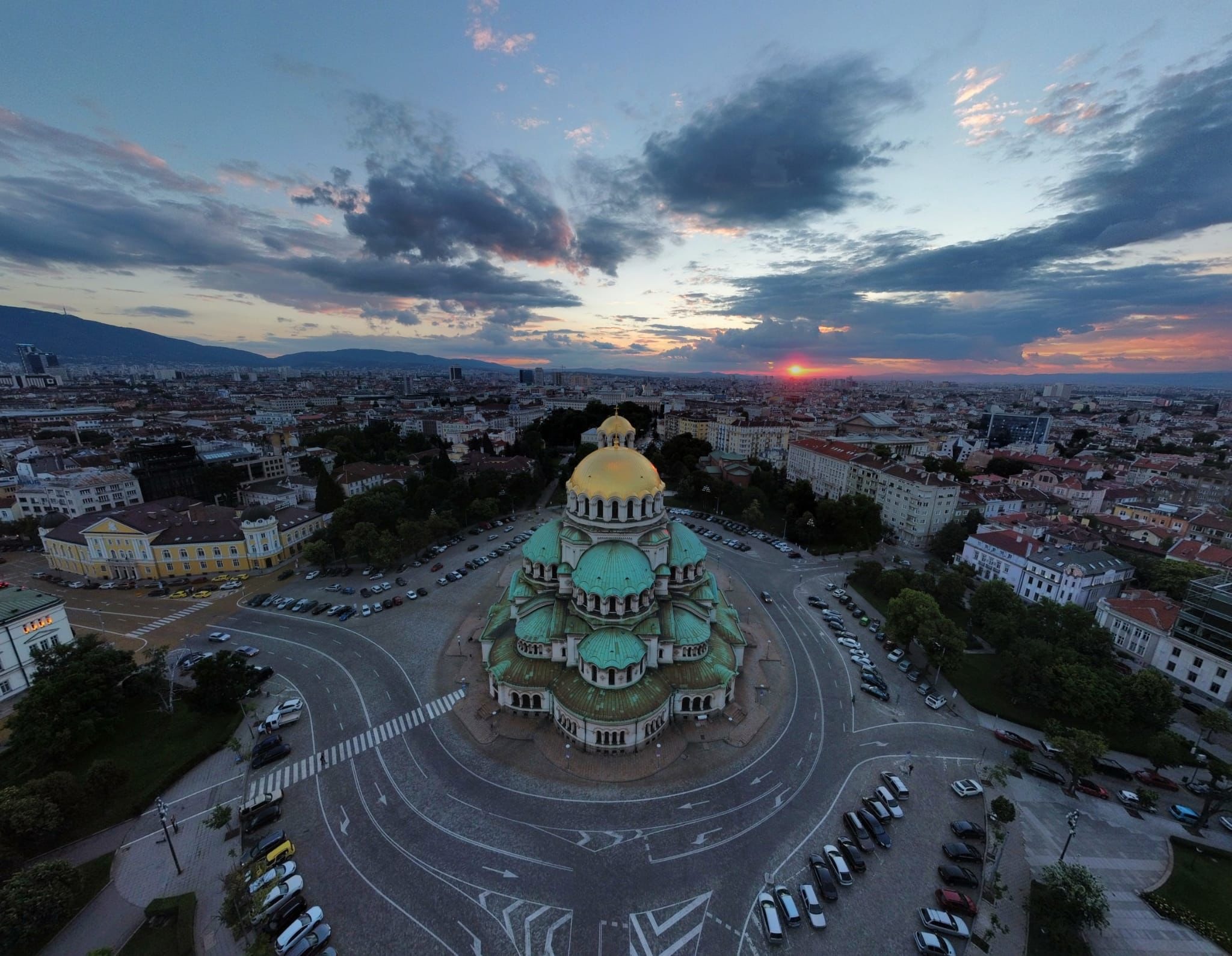Panoramiczny widok z lotu ptaka na sobór z zielonymi kopułami i złotą kopułą główną w Sofii o zachodzie słońca, z widoczną panoramą miasta i górami w tle.