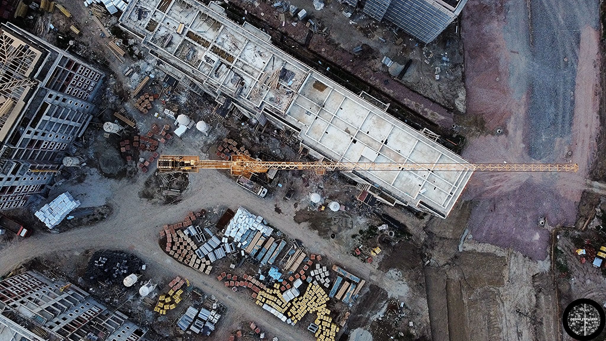 Widok z drona na plac budowy z kilkoma budynkami w stanie surowym, dźwigiem i stertami materiałów budowlanych w różnych kolorach.