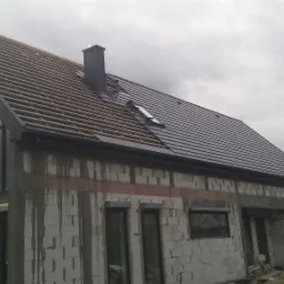 Dom w trakcie budowy z częściowo ukończonym, ciemnoszarym pokryciem dachowym; widoczna konstrukcja drewniana dachu, komin i okno dachowe.