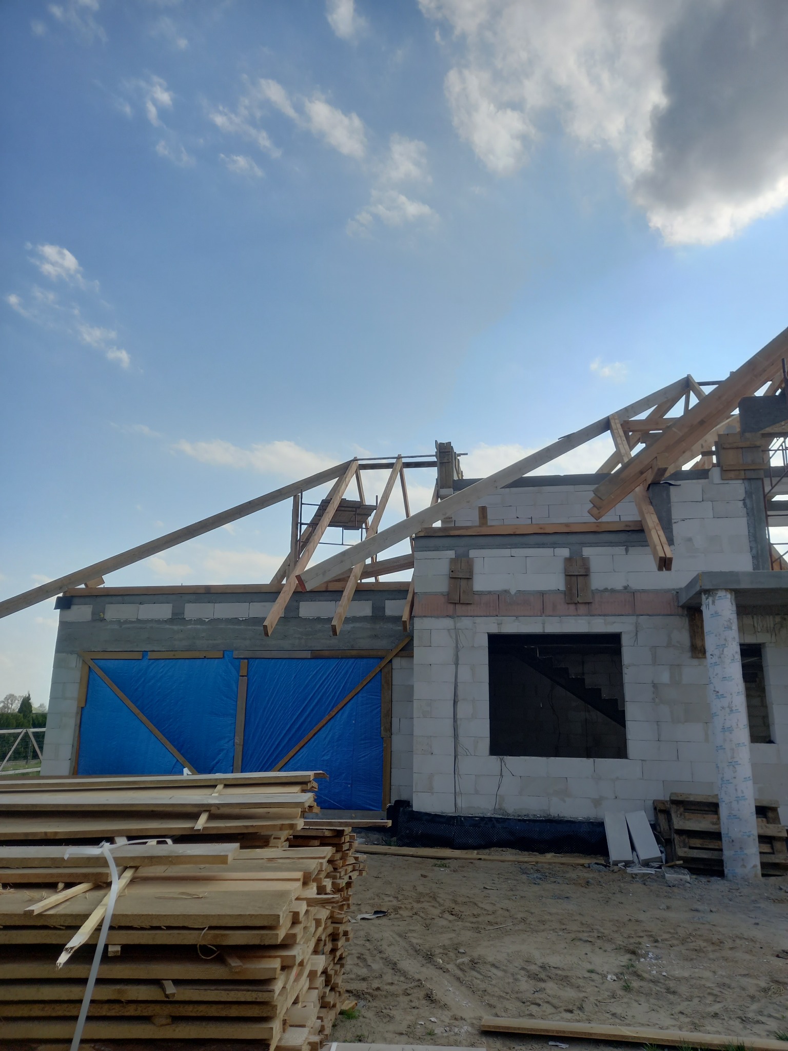 Budowa domu w trakcie wznoszenia konstrukcji dachu; widoczne bale drewniane, ściany z bloczków, niebieska plandeka zabezpieczająca otwory, stos drewna na pierwszym planie, błękitne niebo z chmurami...