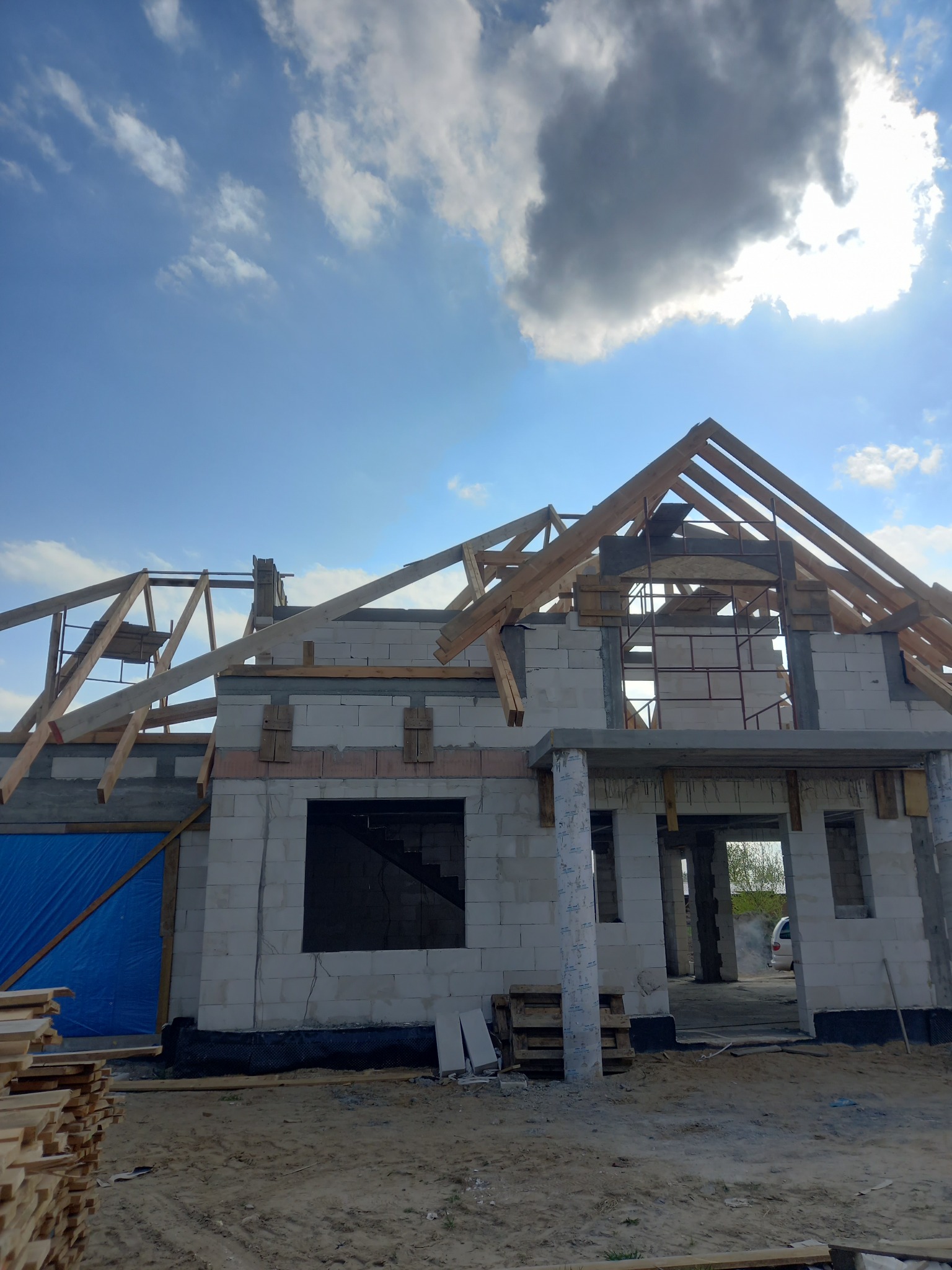 Budowa domu jednorodzinnego w trakcie wznoszenia konstrukcji dachu, widoczne bloczki betonowe, drewniane elementy więźby dachowej oraz niebieska plandeka zabezpieczająca, w tle błękitne niebo...