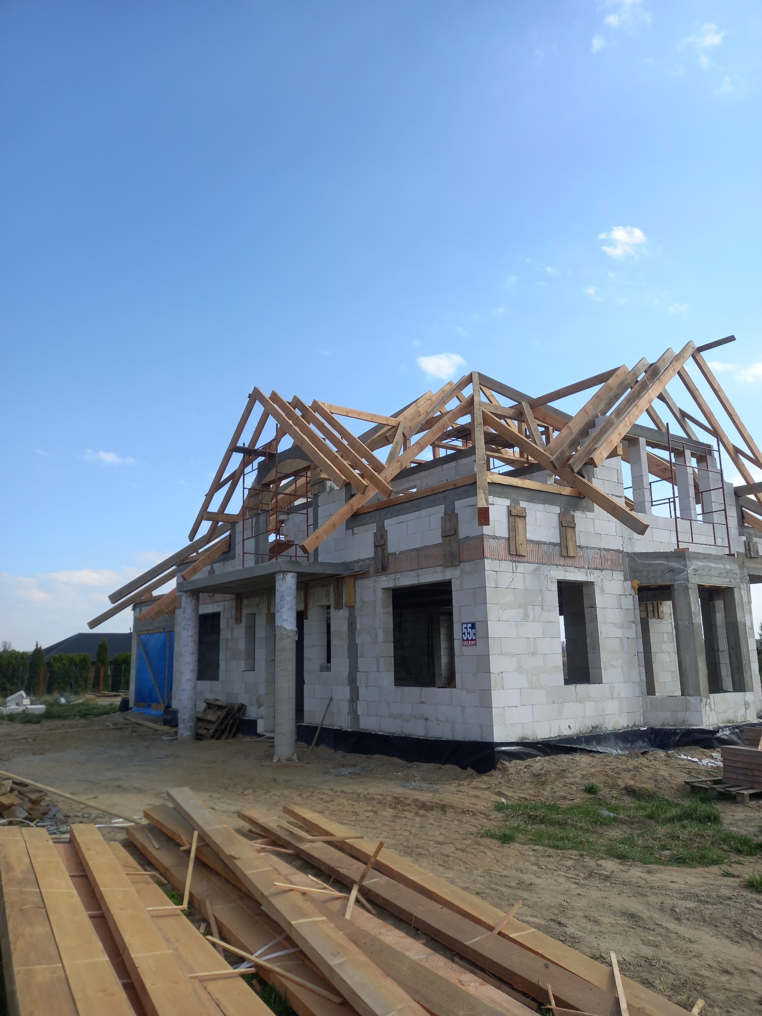 Budowa domu jednorodzinnego w trakcie wznoszenia konstrukcji dachu, widoczne drewniane krokwie i pustaki ceramiczne, na pierwszym planie składowane deski.