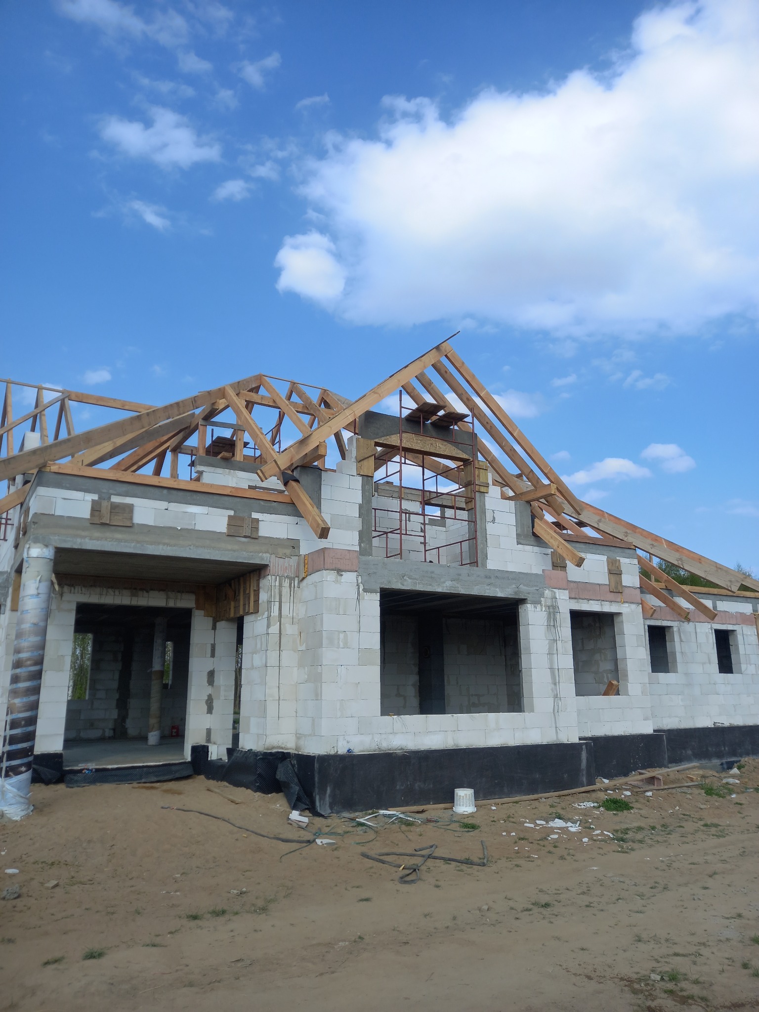 Surowy stan domu jednorodzinnego z pustaków, z widoczną konstrukcją więźby dachowej i elementami deskowania, na tle błękitnego nieba z obłokami.