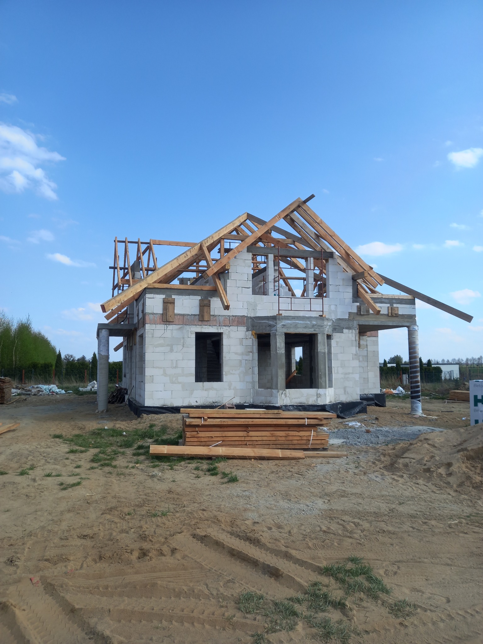 Budowa domu jednorodzinnego w stanie surowym otwartym, widoczne ściany z bloczków, drewniana konstrukcja dachu i stos desek na pierwszym planie.