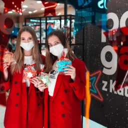Dwie kobiety w czerwonych marynarkach i maseczkach ochronnych trzymają świąteczne pierniki w kształcie gwiazdek przed witryną sklepową Galerii Katowickiej z neonowymi dekoracjami świątecznymi.